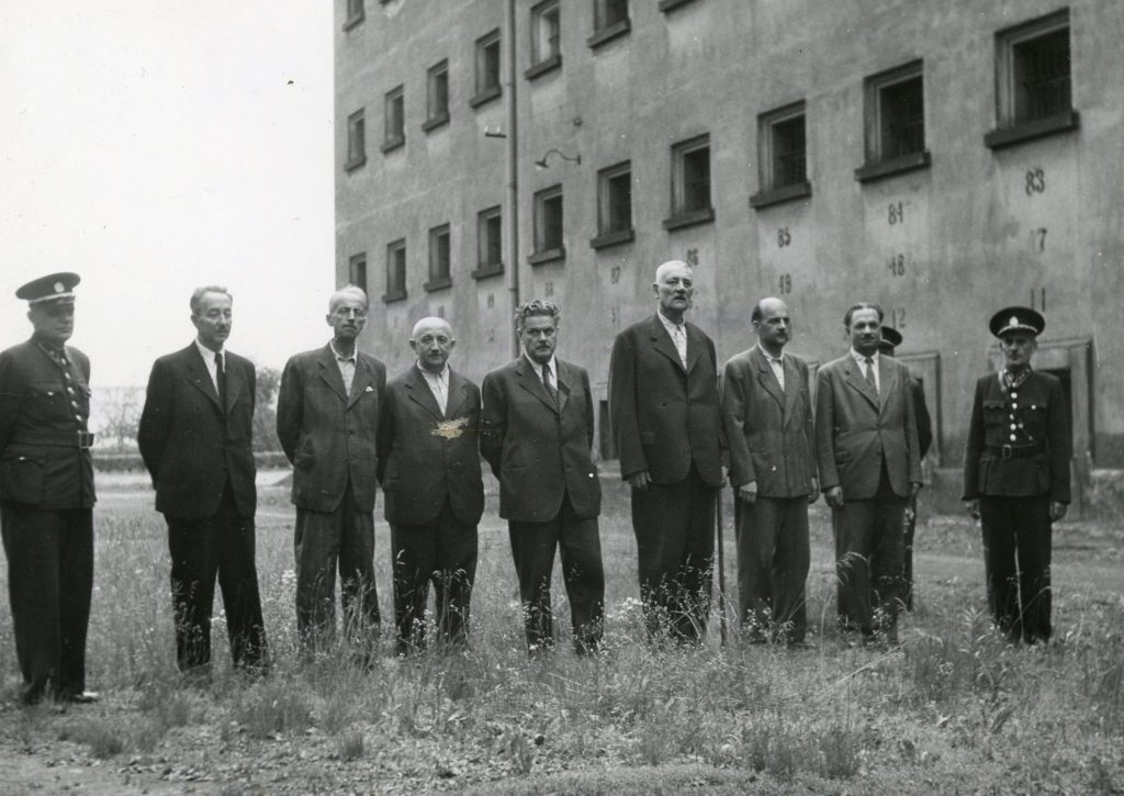 Členové protektorátní vlády na vězeňském dvoře na Pankráci. Mezi dozorci stojí ministři Josef Kalfus, Jindřich Kamenický, Rudolf Beran, Jaroslav Krejčí, Richard Bienert, tajemník Nejvyššího správního soudu a tiskový tajemník prezidentské kanceláře Josef Kliment a ministr Adolf Hrubý.